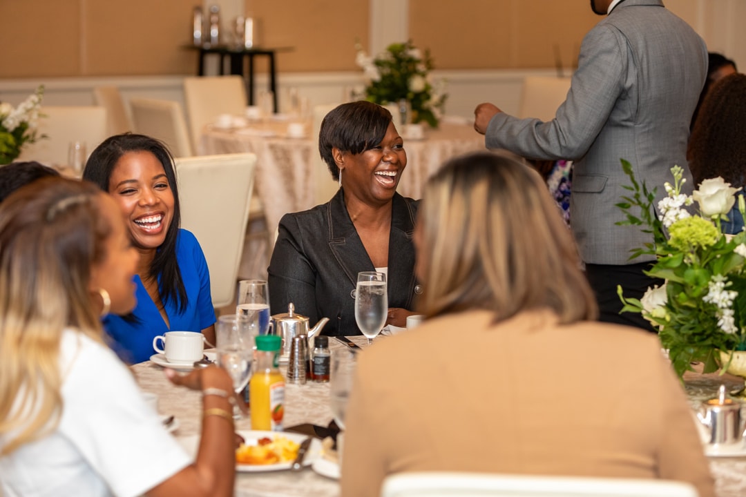Two women at a table laugh joyfully as they engage in discussion, while other attendees and a man in a suit stand in the background. A woman in a mustard-colored sweater sits with her arms crossed, listening intently to a conversation at a round table with a floral centerpiece.