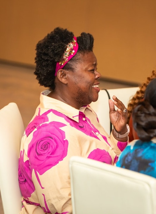 A woman wearing a vibrant floral blouse and a decorative headband laughs while engaged in conversation at a formal event.