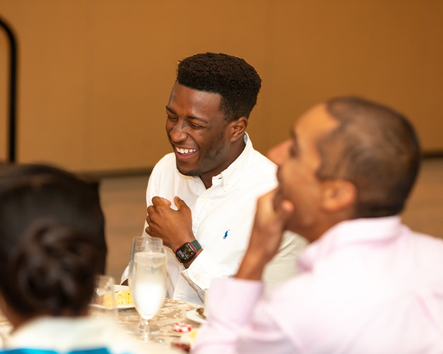 A young man wearing a white shirt and smartwatch laughs while seated at a table, surrounded by other attendees engaged in conversation.