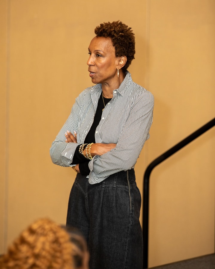 A woman wearing a striped blouse and dark pants, stands with her arms crossed, attentively listening in a professional setting.