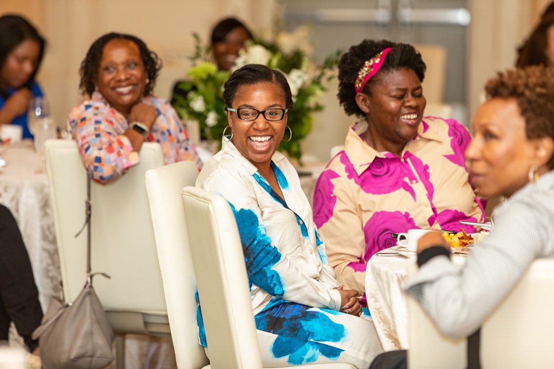 A group of women at a round table laughs and smiles while engaged in conversation, with floral arrangements and plates of food in front of them.