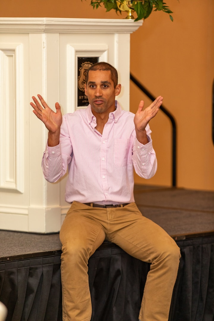 A man in a pink button-up shirt and khaki pants sits on the edge of a stage, gesturing with both hands as he speaks.