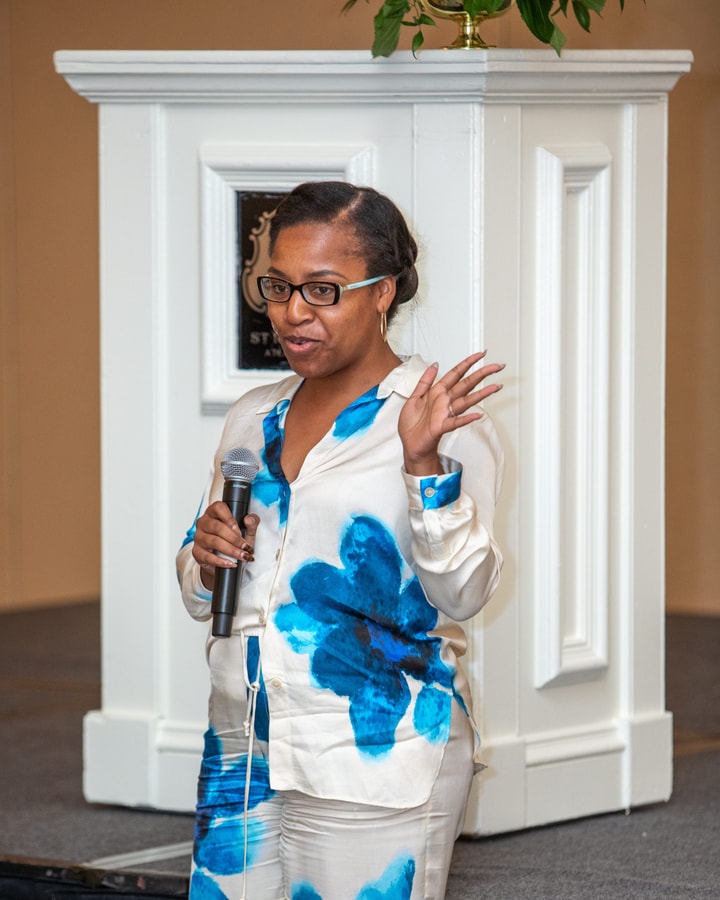 A woman in a white and blue floral outfit holds a microphone while speaking on stage, gesturing with one hand in front of a white podium.