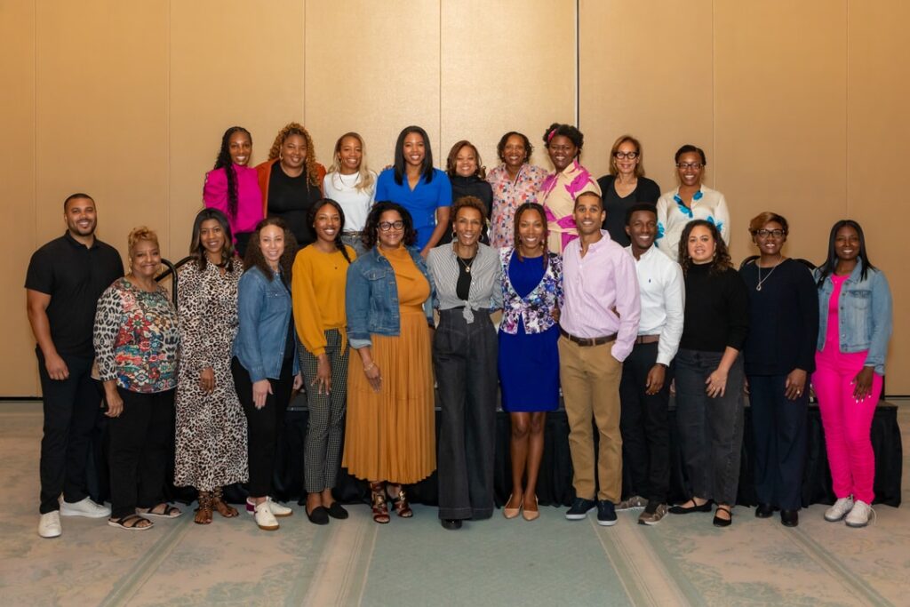 A group of people poses for a photo in a conference or event setting. They are dressed in a mix of business casual and formal attire, standing in two rows in front of a beige wall.