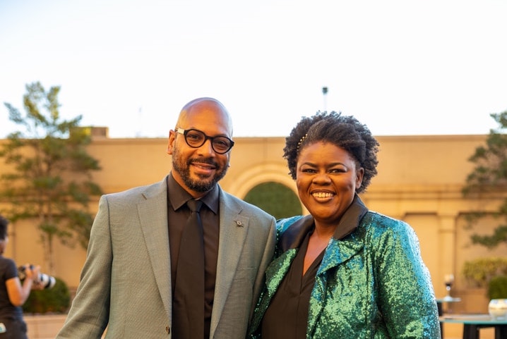 An elegantly dressed man and woman smiling and posing for a photo outdoors.