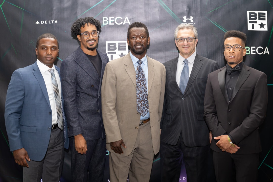 Five men, dressed in suits of varying colors, stand side by side and smile at the camera in front of a sponsor-branded backdrop.