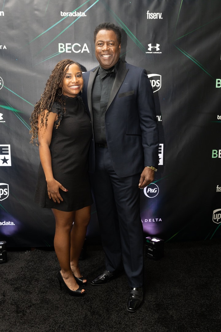 A man in a dark suit and bow tie stands beside a woman in a short black dress, both smiling against a sponsor-branded backdrop.