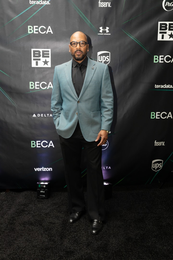 A man wearing a light blue blazer, black shirt, and black tie stands in front of a sponsor-branded backdrop.