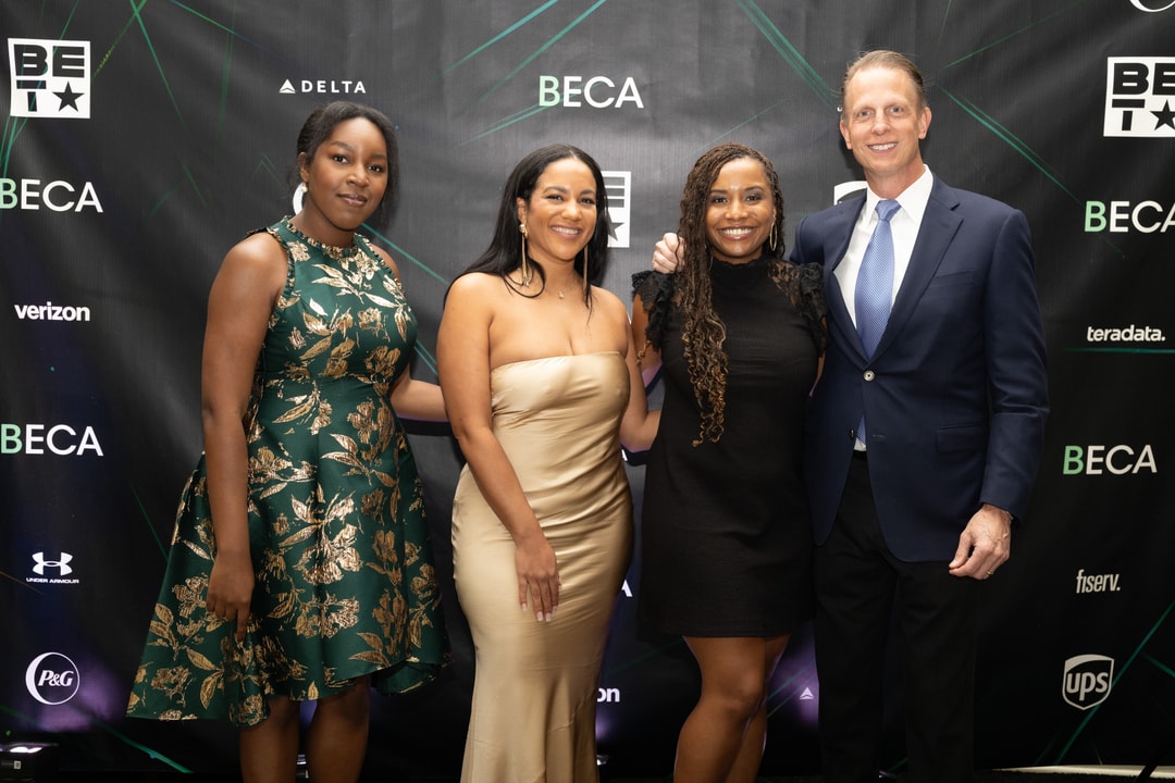 Four individuals pose together, smiling, in front of a sponsor-branded backdrop. Three women wear dresses in green, gold, and black, while the man stands at the right in a navy suit.