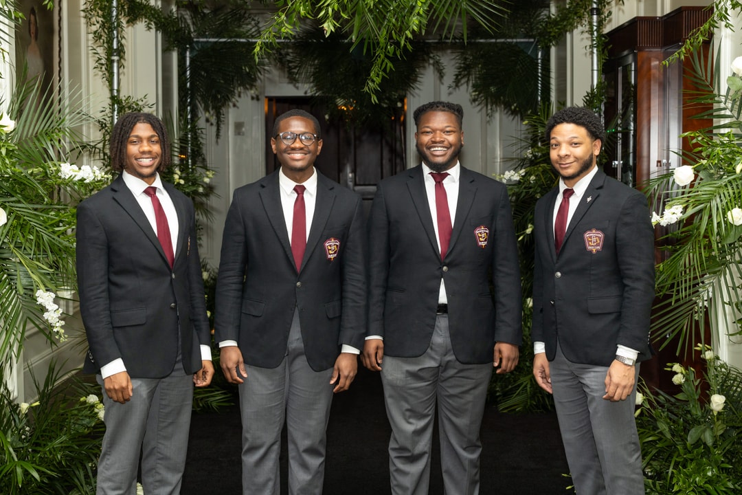 Four young men in matching black blazers with embroidered crests, white shirts, and burgundy ties stand together, smiling, in a hallway decorated with lush green foliage and white flowers.