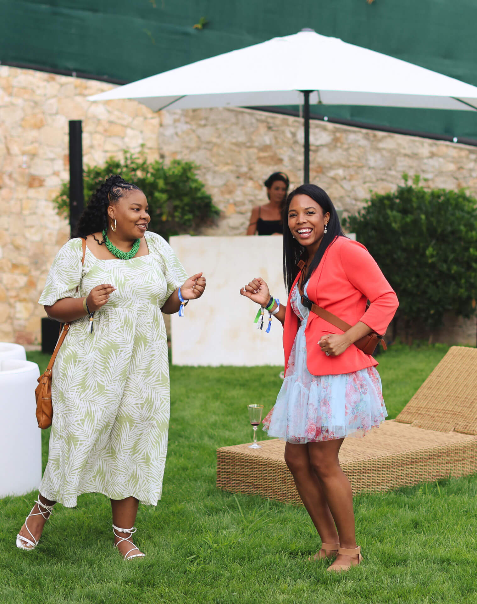 Two women in summery outfits, smiling and dancing on a lawn with a lounge chair and umbrella in the background.