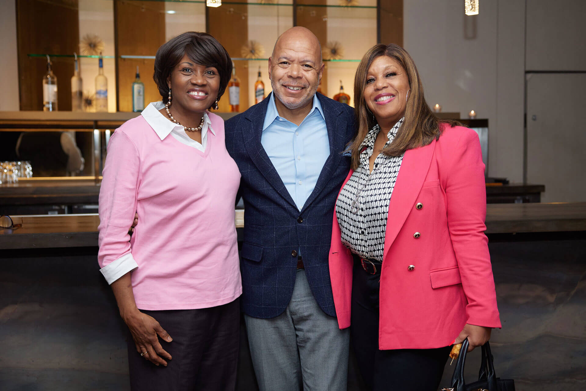 Three individuals standing side by side, smiling in front of a bar setup.