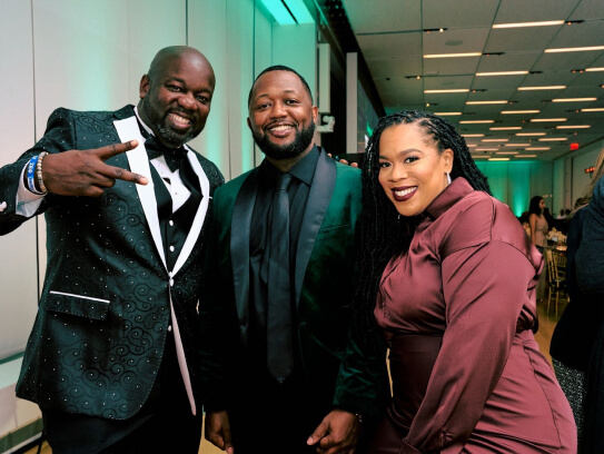Three attendees dressed in formalwear posing for a photo, two men in tuxedos and a woman in a burgundy dress.