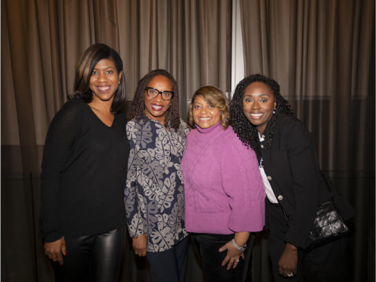A group of four women standing close together and smiling at the camera in front of a draped backdrop.
