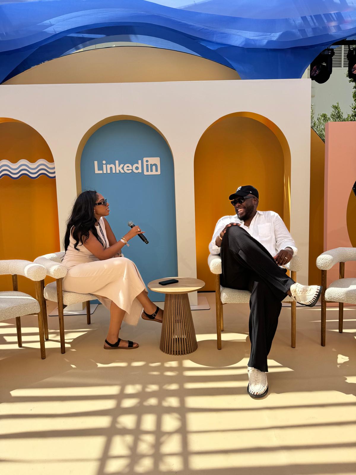 A woman and a man seated on a stage with a LinkedIn backdrop, engaged in a conversation. The woman holds a microphone while the man, wearing sunglasses and a hat, smiles as they talk.
