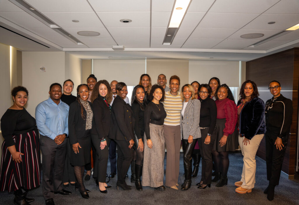 A group of professionals in business attire stands in a meeting room, smiling as they pose for a group photo.
