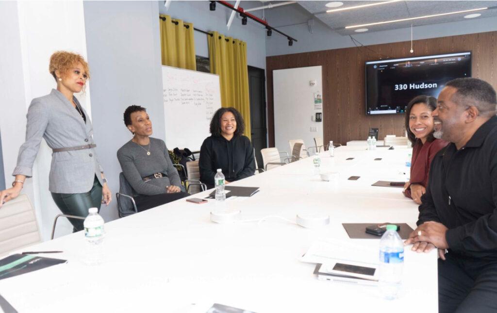 A small group of colleagues sits around a long conference table in a modern office, smiling and talking as one woman stands at the head of the table. A screen in the background displays â330 Hudson.â