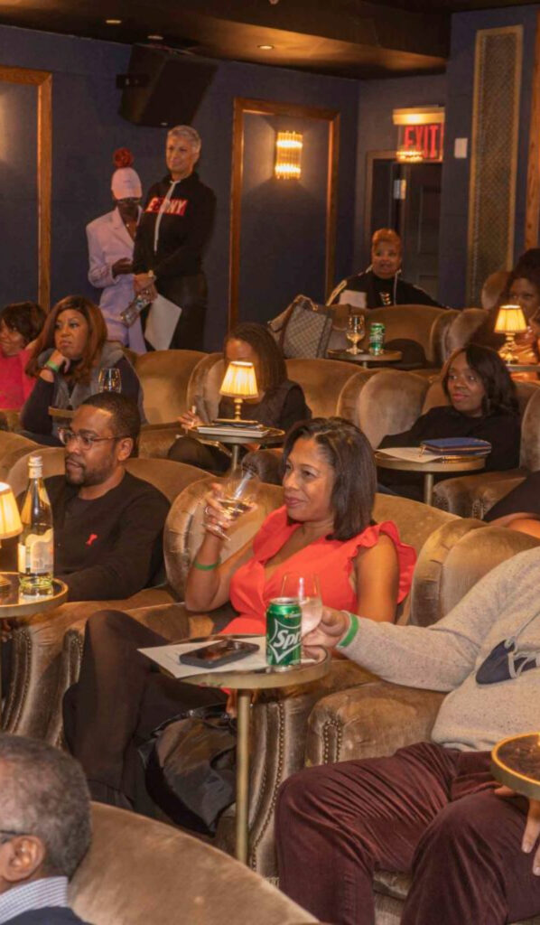 Audience members sit in plush lounge chairs, sipping drinks and watching an event in a dimly lit room, while a few people stand near the back.