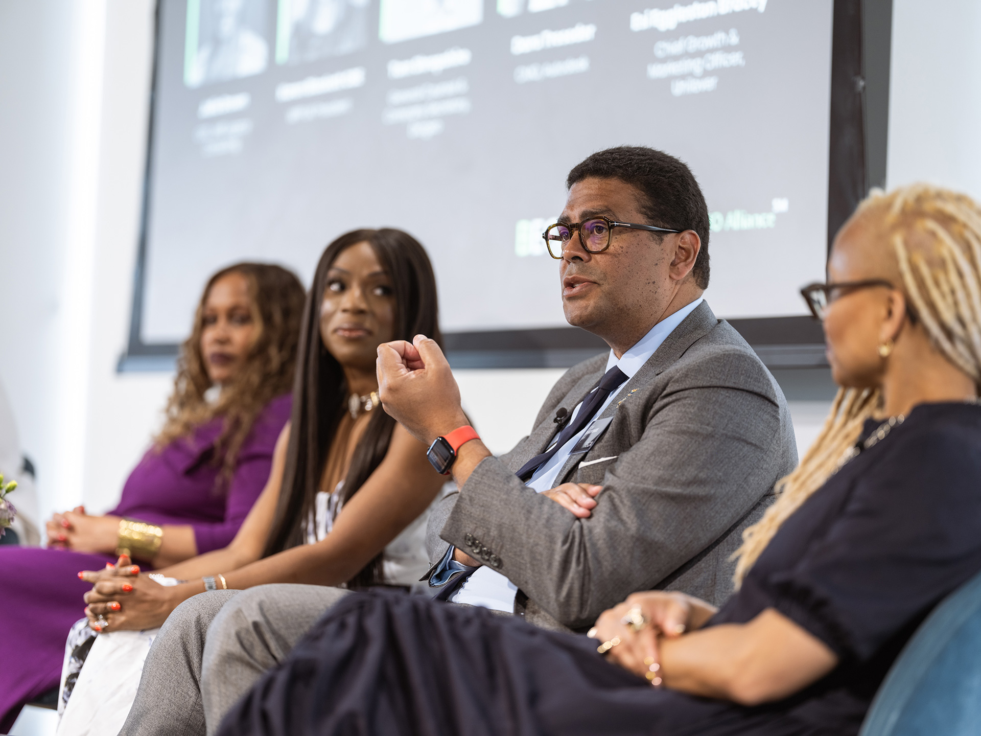 Four panelists seated in a row, with one male speaker in a gray suit talking while the three female panelists listen attentively.