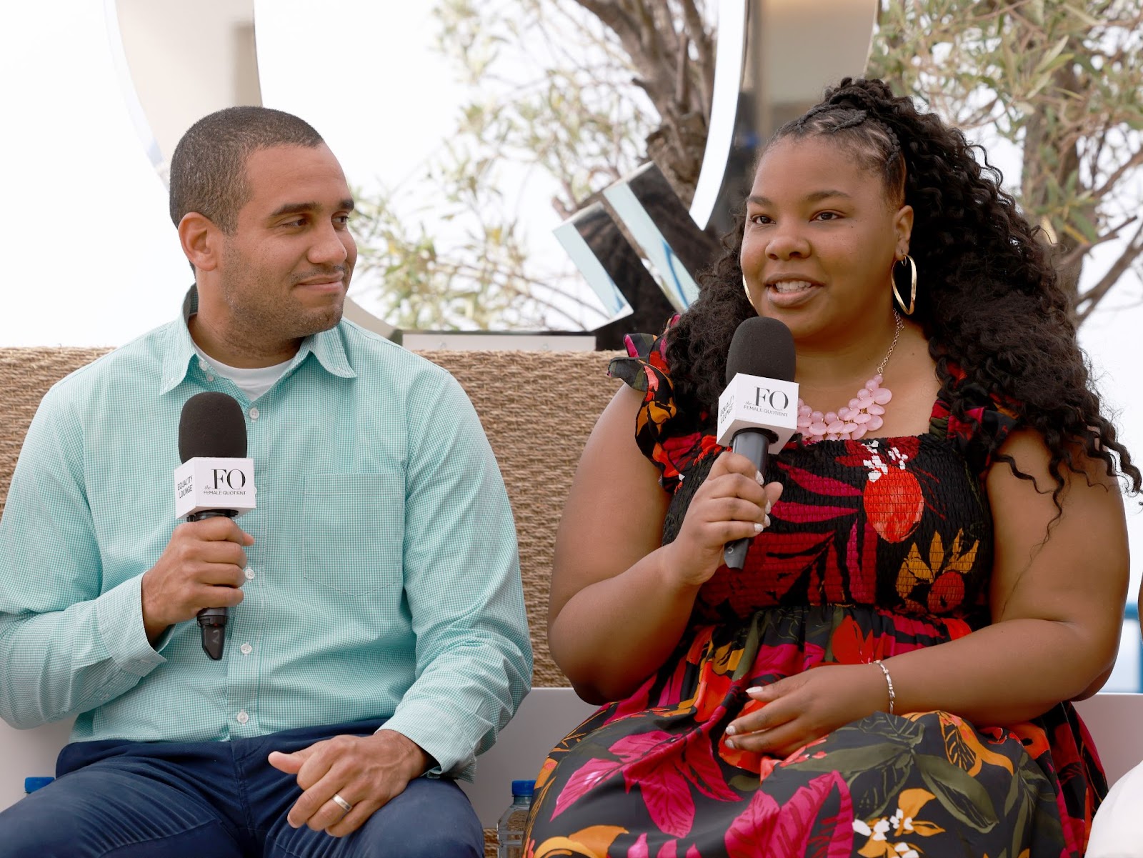 A man in a light green shirt and a woman in a floral black and red dress, both holding microphones and engaged in a conversation during an event.