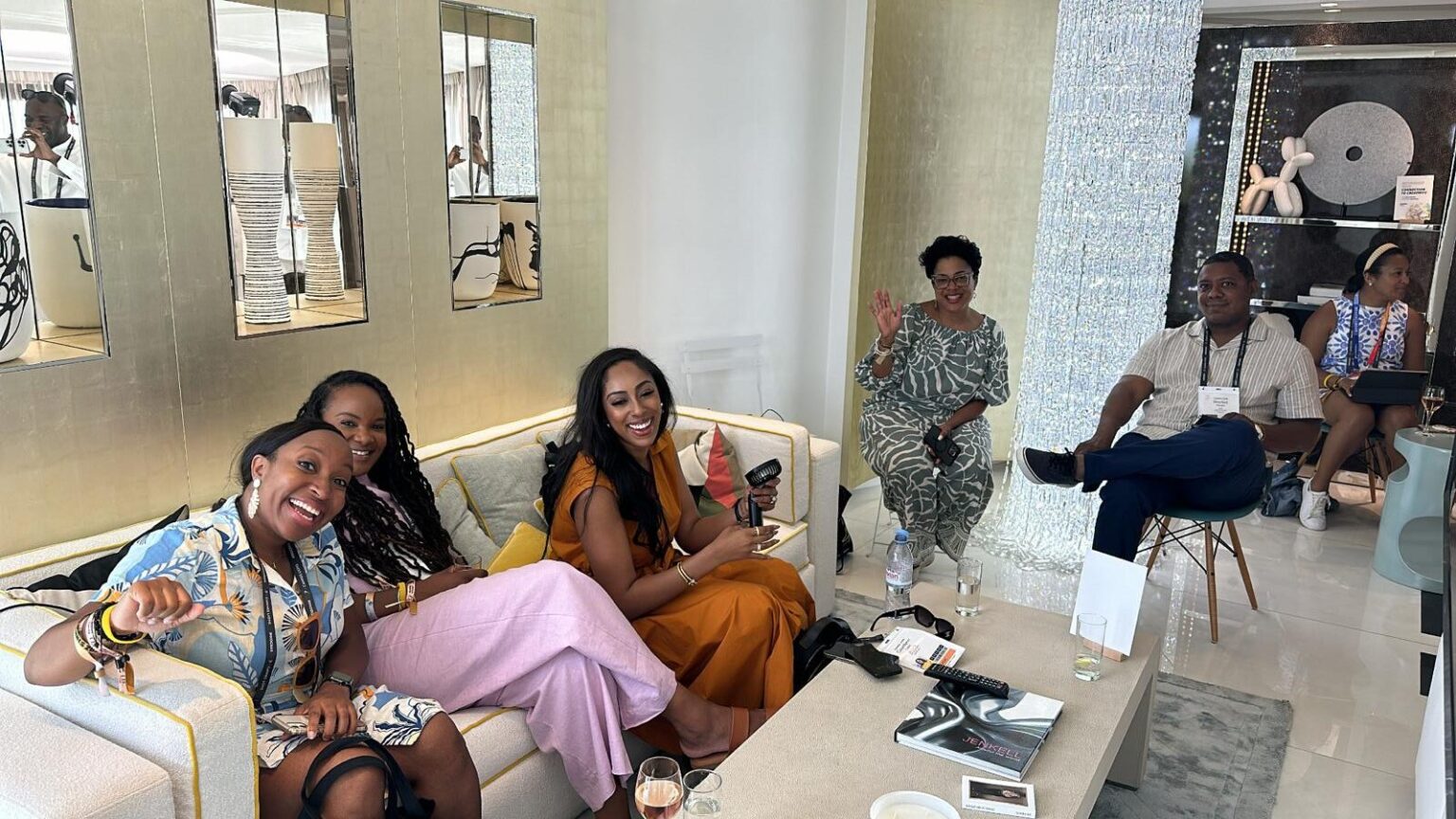 A group of six people is gathered in a stylish, modern lounge with gold accents, mirrors, and decorative art pieces. They are seated on plush furniture, smiling, chatting, and enjoying the relaxed atmosphere. Some are holding drinks, while others wave at the camera. A well-lit and elegant interior design sets the scene, with reflections of additional people visible in the mirrors.
