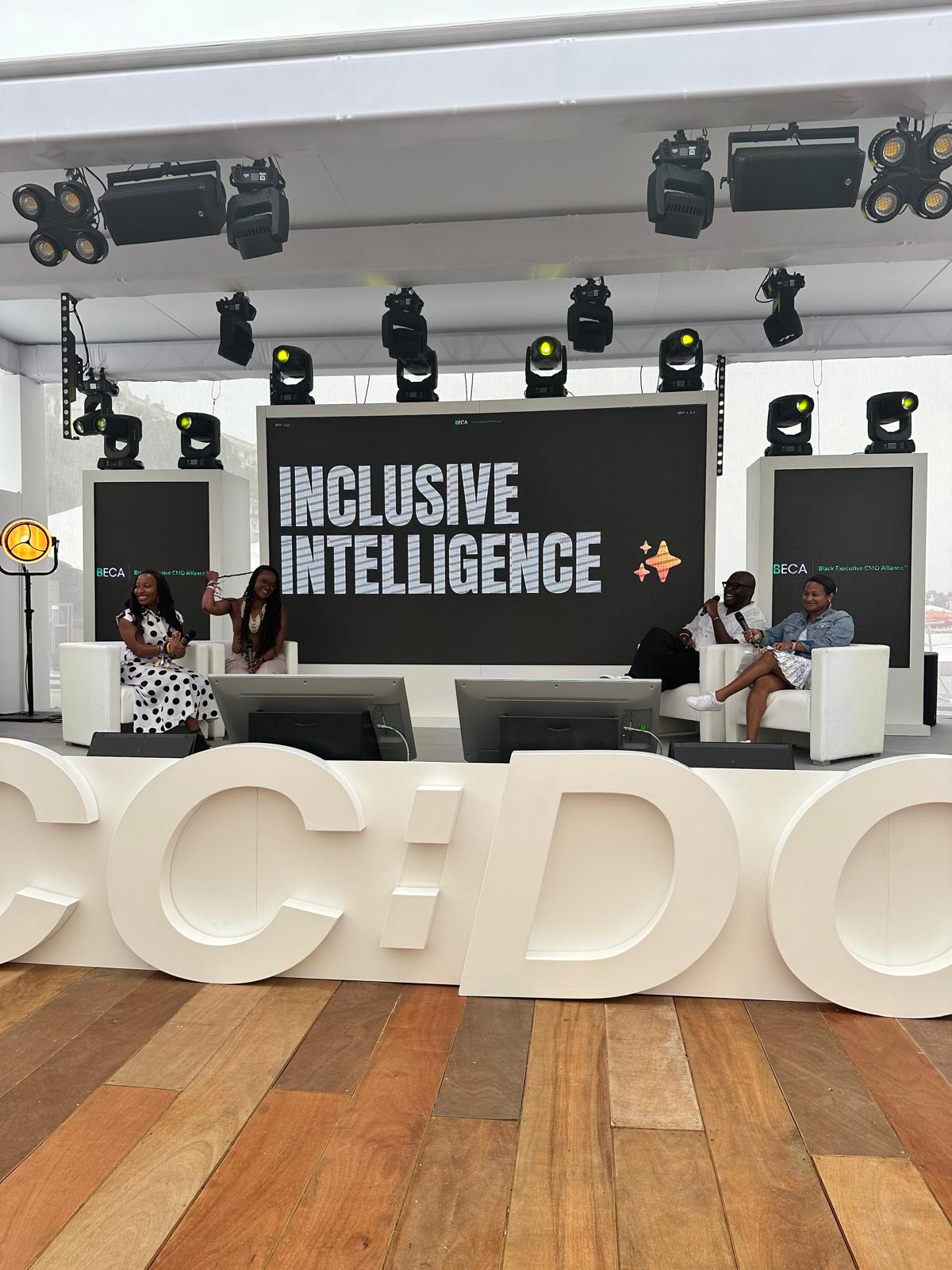 A panel discussion is taking place on a professionally lit stage at a BECA (Black Executive CMO Alliance) event. The large screen behind the speakers displays the words "INCLUSIVE INTELLIGENCE." Four individuals are seated on modern white chairs, actively engaging in the discussion. The stage features sleek lighting, multiple screens, and bold white letters in the foreground, contributing to a polished and dynamic event setting.