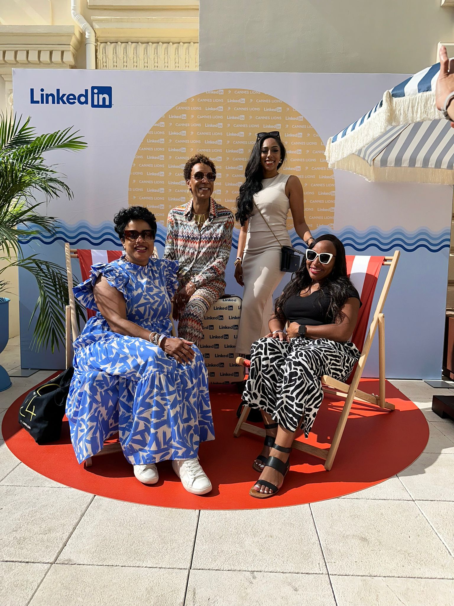 Four women pose for a photo at a LinkedIn-branded event space with a beach-themed backdrop featuring a sun and waves. Two women are seated in red-and-white striped deck chairs, while the other two stand behind them. They are dressed in stylish summer attire, with one wearing a blue patterned dress, another in a patterned jumpsuit, a third in a beige fitted dress, and the fourth in a black-and-white patterned skirt with sunglasses. The setting is outdoors with a blue-and-white striped umbrella providing shade.