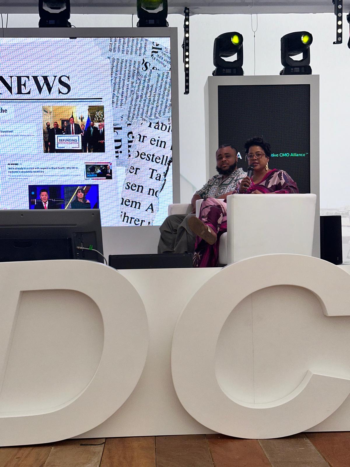 Two panelists are seated on a stage at a BECA (Black Executive CMO Alliance) event, engaged in a discussion. The woman, wearing a patterned outfit, is speaking into a microphone, while the man listens attentively. Behind them, a large screen displays a collage of newspaper clippings and news headlines, including an image with the word "DEFUNDING." The stage is professionally set up with bright lighting, modern design elements, and large white letters in the foreground.