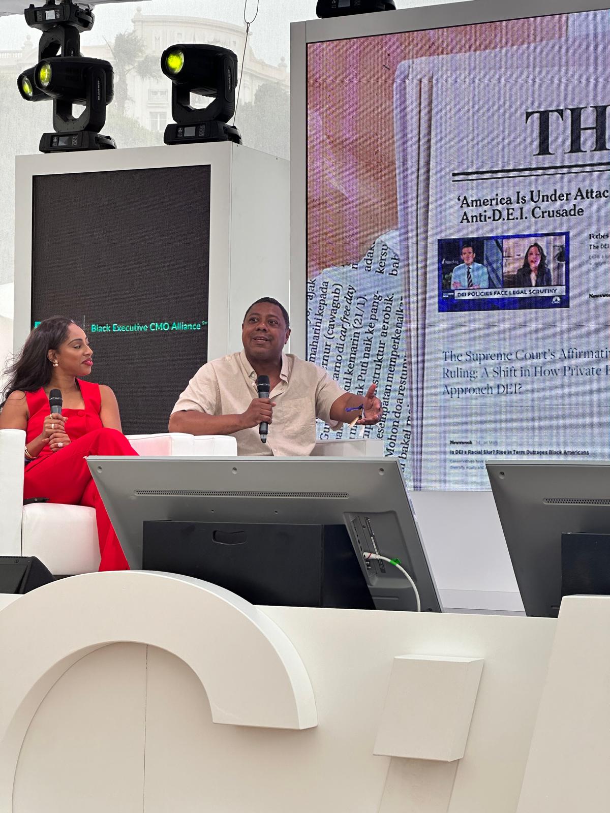 A man and a woman are seated on a stage at a BECA (Black Executive CMO Alliance) event, engaging in a discussion. The man is speaking into a microphone while gesturing, and the woman in a red outfit listens attentively. Behind them, a large screen displays a newspaper article with the headline "America Is Under Attack: Anti-D.E.I. Crusade," along with an image of a news broadcast discussing DEI policies. The stage is professionally lit, featuring modern design elements and large white letters in the foreground.