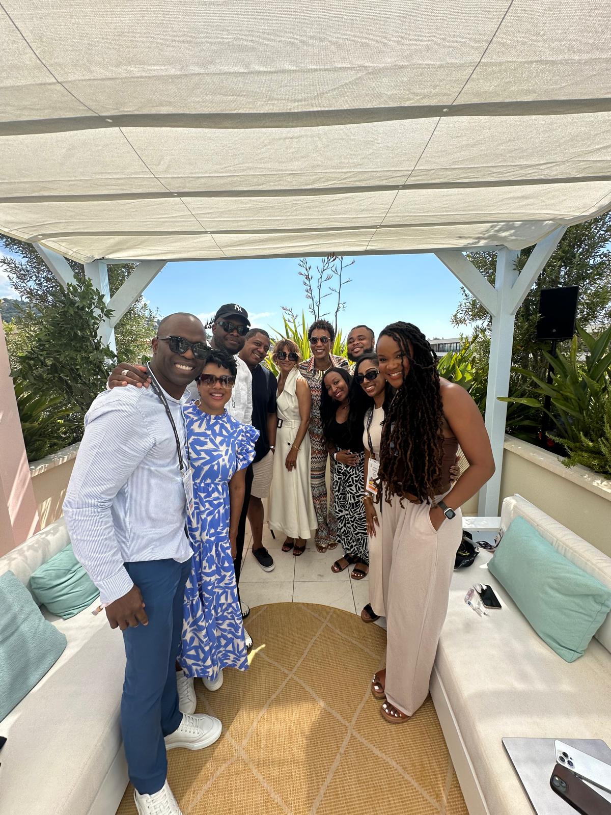 A group of ten diverse individuals pose together under a shaded outdoor pergola, smiling at the camera. They are dressed in stylish, summery outfits, with some wearing sunglasses. The setting features lush greenery, comfortable seating, and a bright blue sky in the background, suggesting a relaxed and elegant event.