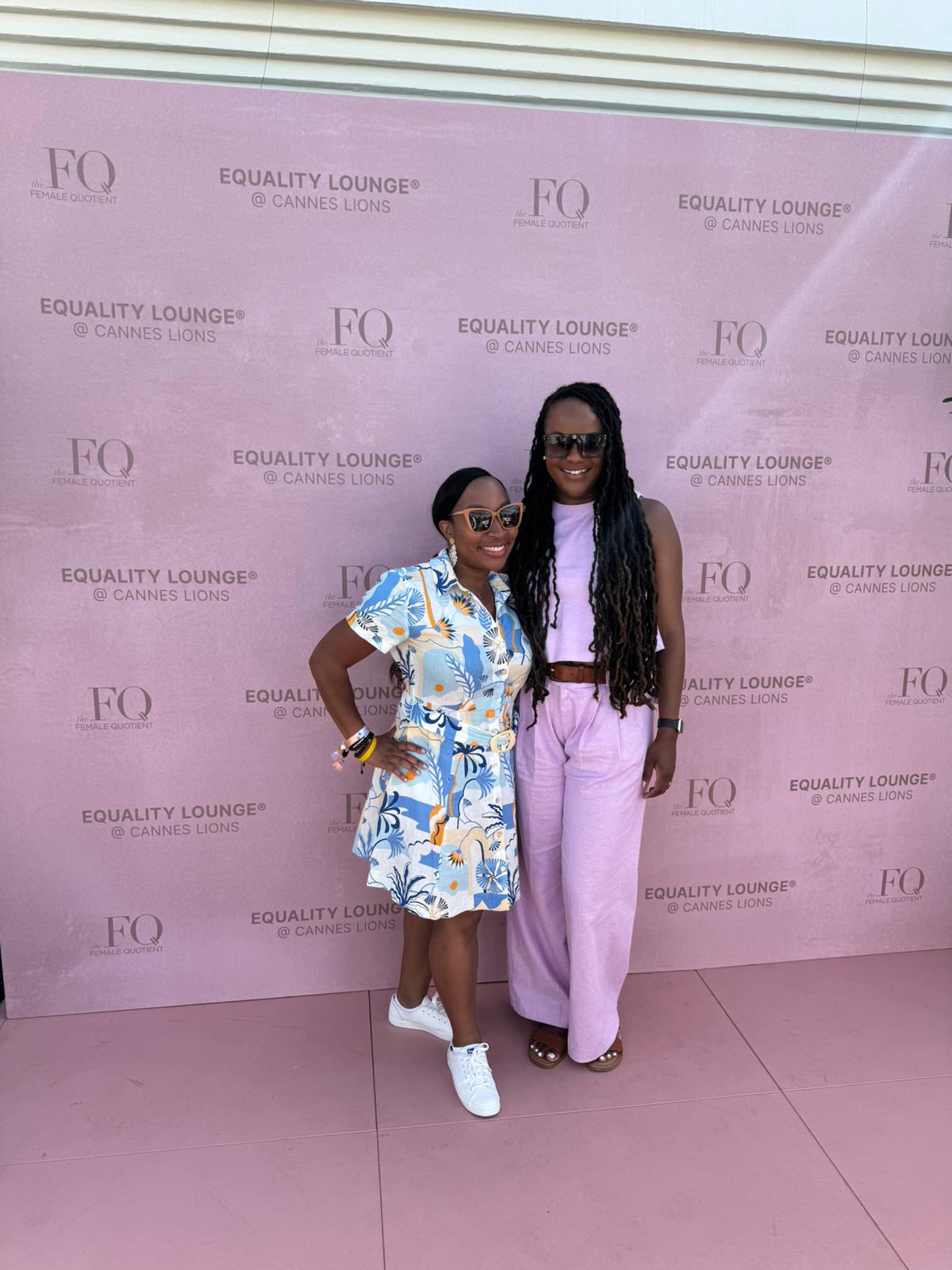 Two women pose together in front of a pink backdrop featuring the branding for "Equality Lounge @ Cannes Lions" and The Female Quotient (FQ). They are smiling and dressed in summery outfits—one in a floral print dress with white sneakers and the other in a lavender pants and top set with sandals. Both wear sunglasses, adding to the stylish and relaxed atmosphere of the event.