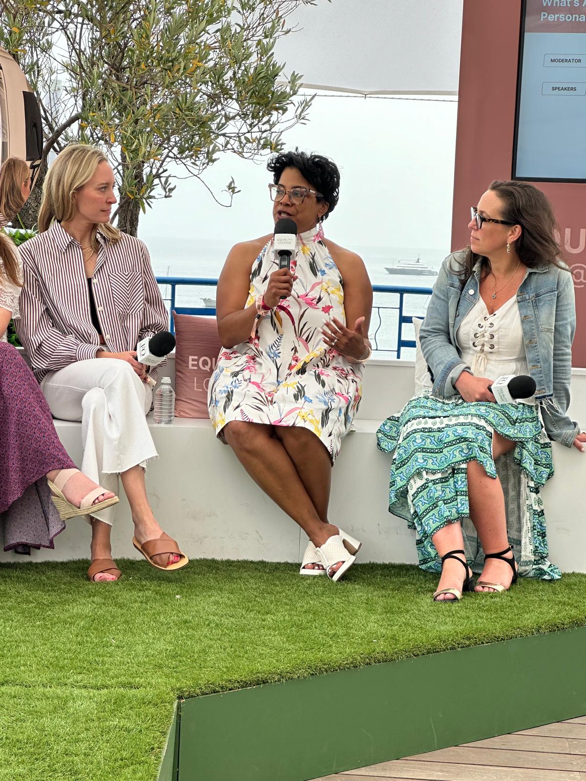 Three women are seated on an outdoor stage, engaged in a panel discussion at an Equality Lounge event. The woman in the center, wearing a floral halter dress, is speaking into a microphone while the other two, dressed in stylish casual outfits, listen attentively. The backdrop features a scenic ocean view with a yacht in the distance, and a screen displaying event details. The setting is relaxed yet professional, with a focus on meaningful conversation.