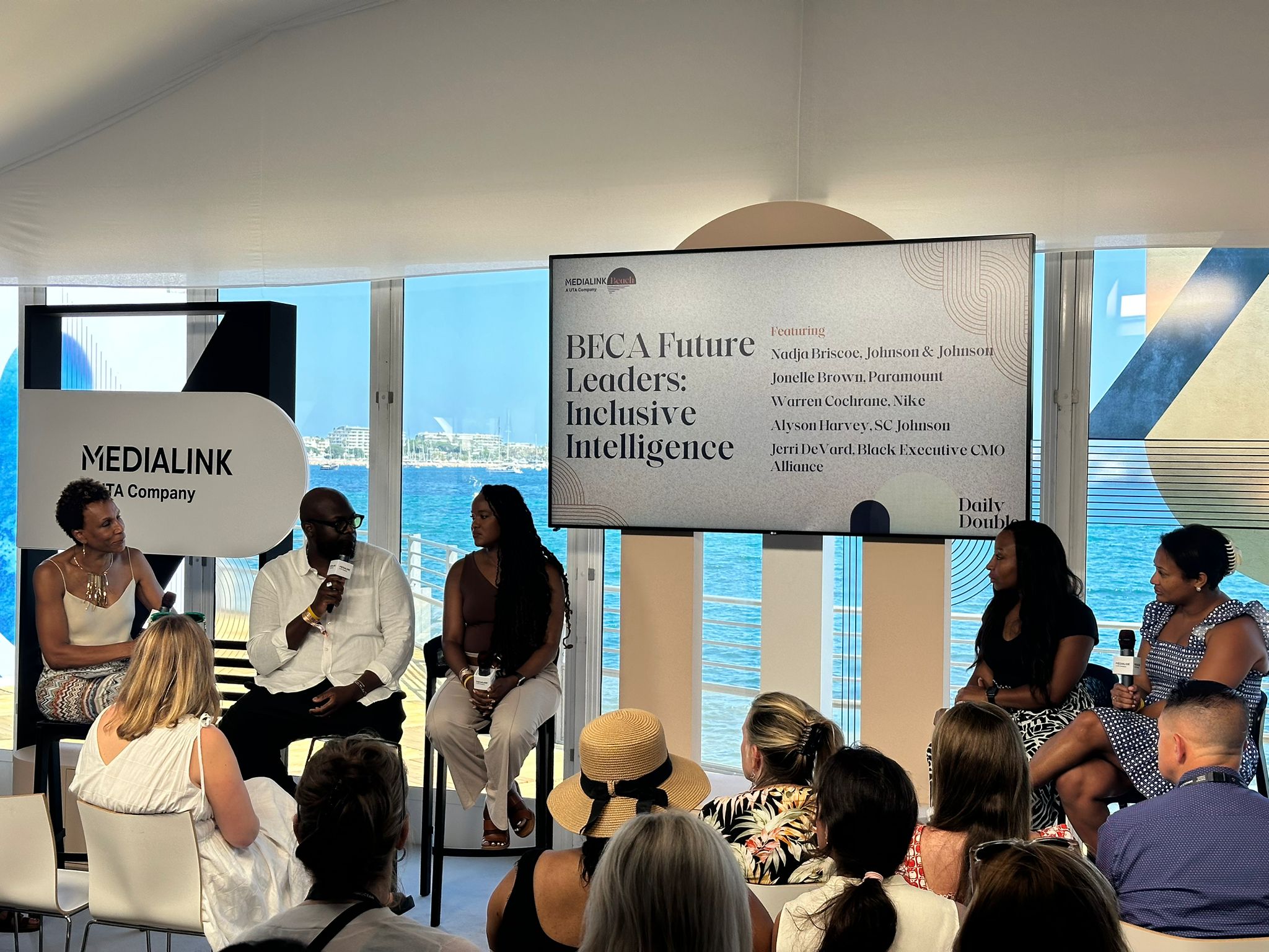 A panel discussion titled "BECA Future Leaders: Inclusive Intelligence" takes place in a bright, modern venue with a view of the ocean in the background. Five panelists, including men and women, are seated on stools, engaged in conversation. The large screen behind them displays the event title and names of featured speakers from companies such as Johnson & Johnson, Paramount, Nike, SC Johnson, and the Black Executive CMO Alliance. The audience, dressed in summer attire, listens attentively. The event is hosted by MediaLink, as indicated by the signage.