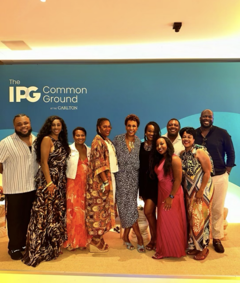 A diverse group of individuals posing together in front of a blue wall with the text "The IPG Common Ground at the Carlton," smiling at an indoor event.