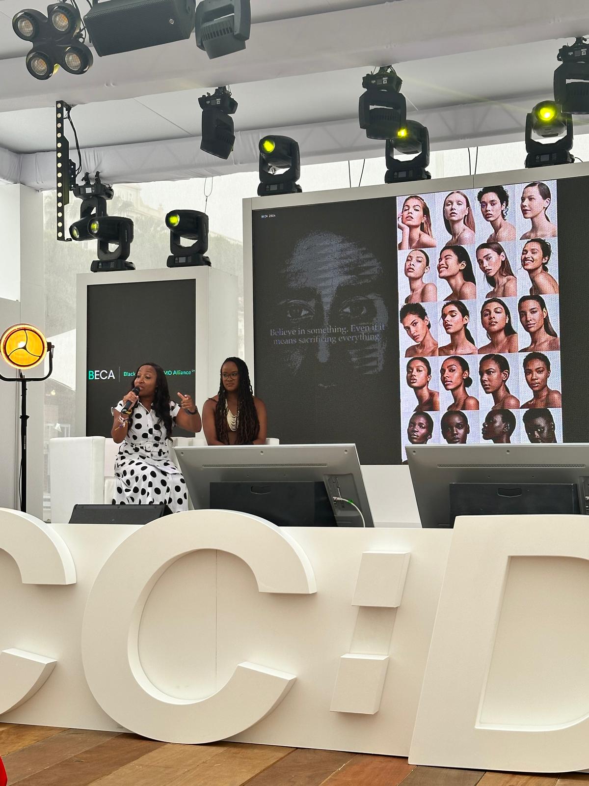 Two women are presenting on a stage at a BECA (Black Executive CMO Alliance) event. One is speaking into a microphone while the other listens attentively. A large screen behind them displays a Nike "Believe in Something" ad featuring Colin Kaepernick and a grid of diverse female portraits. The stage is professionally lit, with modern design elements, including large white letters in the foreground.