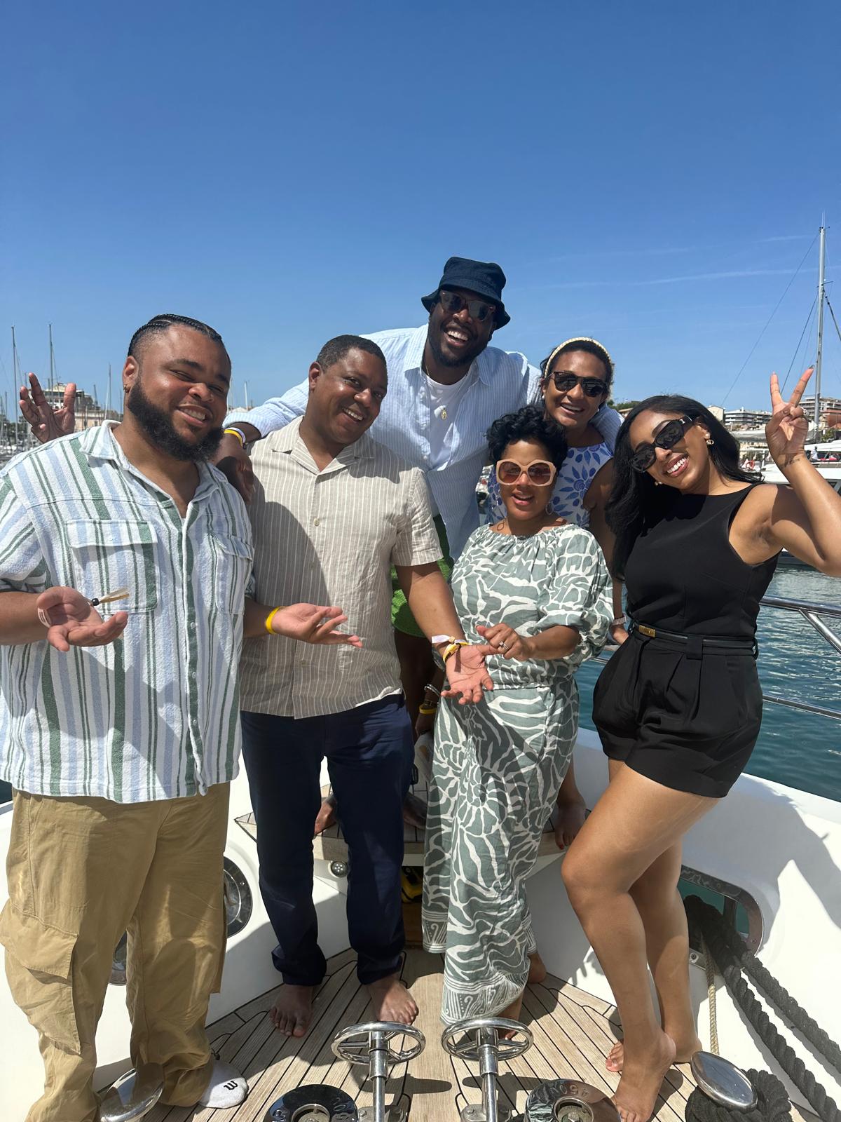 A group of six people poses playfully on the deck of a yacht under a bright blue sky, with a marina and cityscape in the background. They are dressed in stylish, summery outfits, wearing sunglasses, and making fun gestures, including peace signs and shrugging expressions. The relaxed and cheerful mood suggests they are enjoying a day on the water.