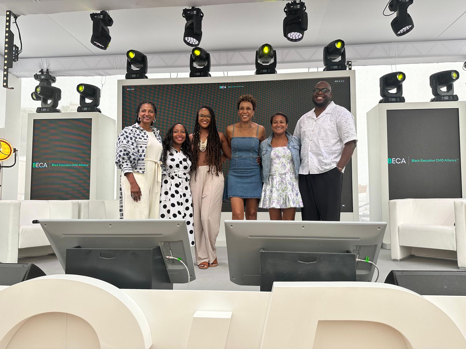 A group of five people stands on a well-lit stage with a modern setup, smiling at the camera. The stage features large screens displaying "BECA" and "Black Executive CMO Alliance," along with professional lighting and white seating. The individuals are dressed stylishly in a mix of casual and semi-formal attire, indicating a professional yet welcoming event atmosphere.