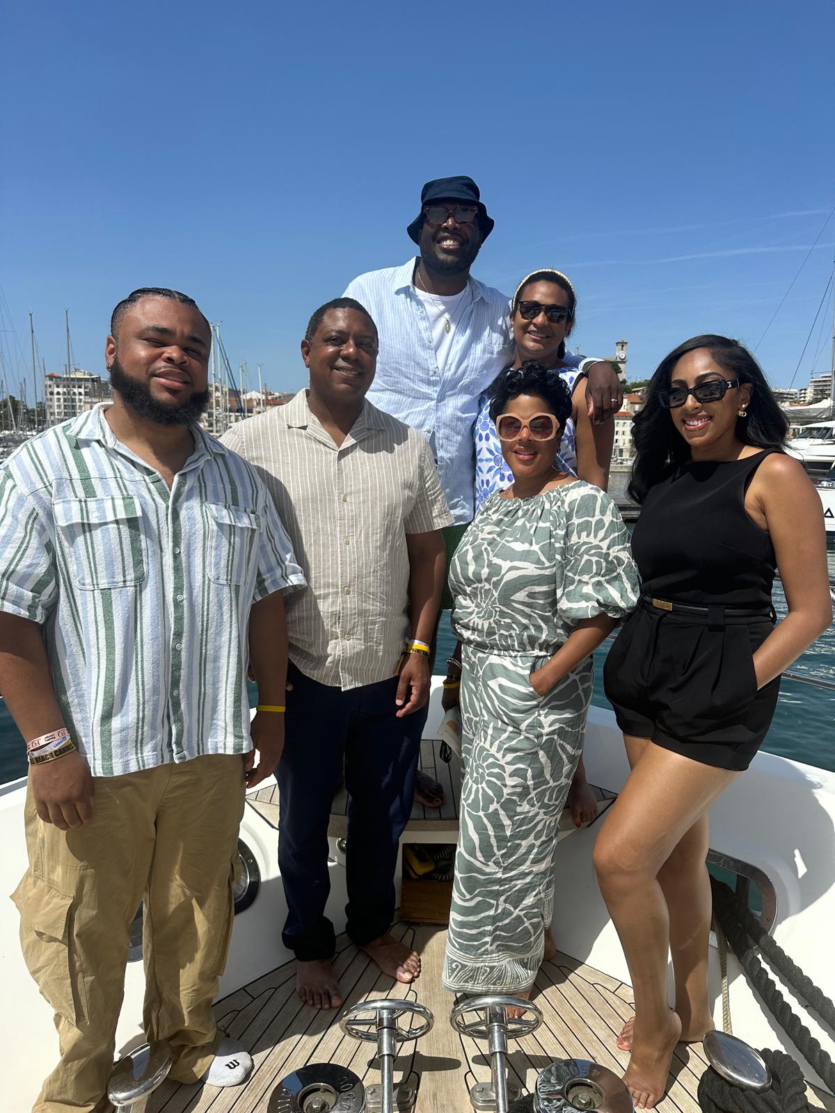 A group of six people poses on the deck of a yacht under a clear blue sky, with a marina and cityscape in the background. They are dressed in stylish, summery outfits, wearing sunglasses and smiling. The relaxed and elegant setting suggests a luxurious day on the water.
