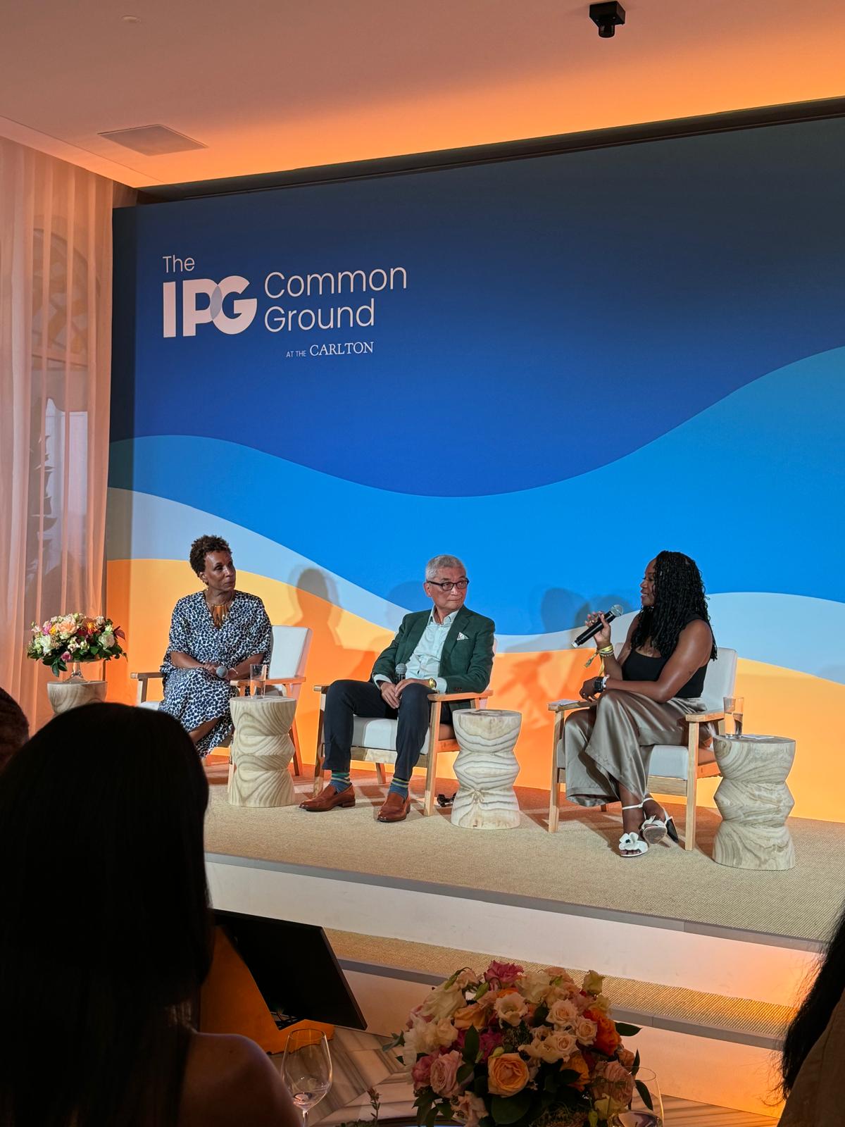 Three panelists are seated on a stage at The IPG Common Ground at The Carlton event, engaged in a discussion. The stage has a blue and orange wave-themed backdrop, with modern wooden stools and floral arrangements. One woman is speaking into a microphone while the other two panelists, a woman in a patterned dress and a man in a green blazer, listen attentively. The setting suggests a professional yet inviting atmosphere for conversation and thought leadership.