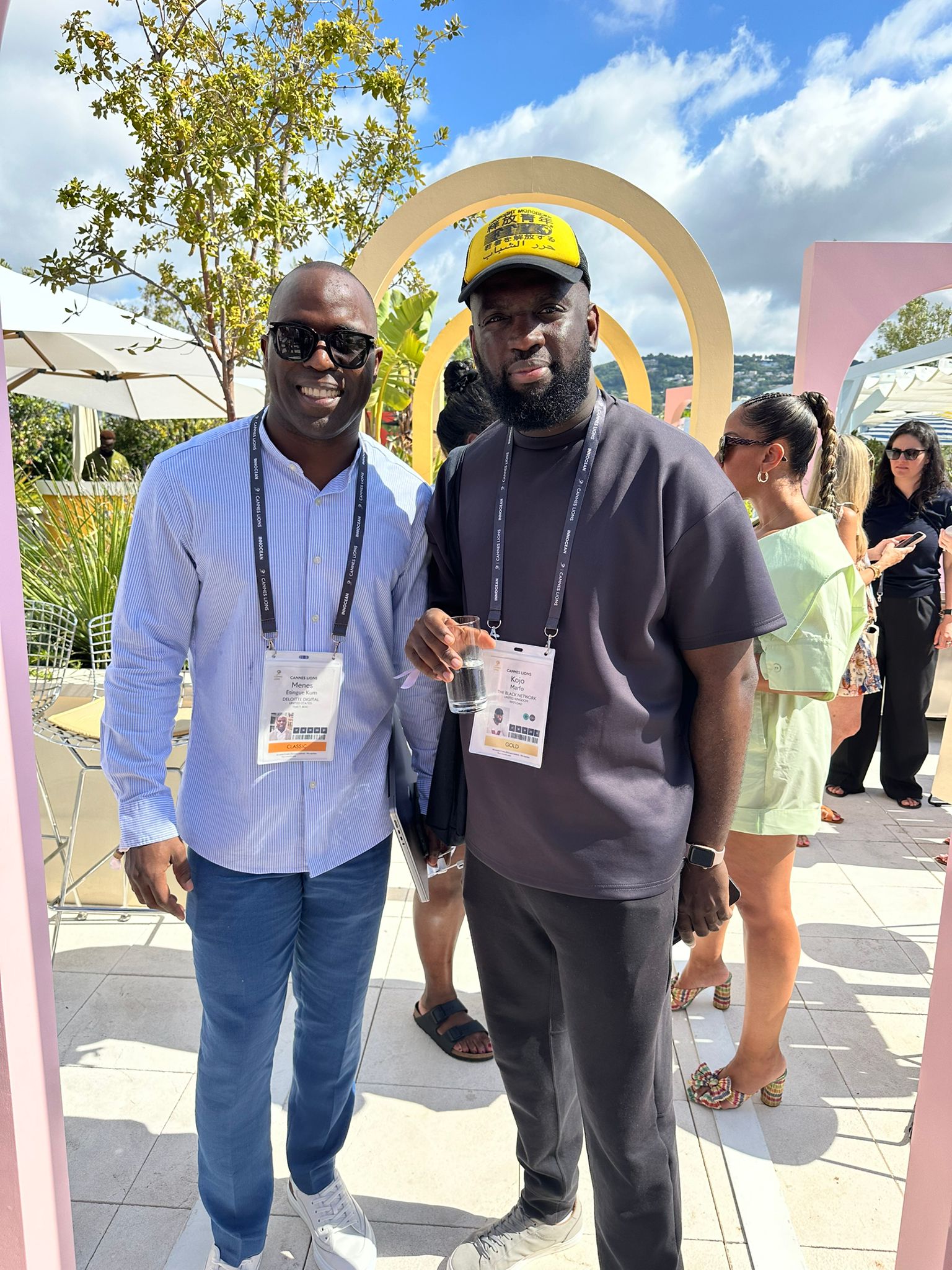 Two men pose for a photo at an outdoor networking event under a sunny sky. One wears a light blue button-up shirt, sunglasses, and a lanyard with a name badge, while the other wears a black t-shirt, a yellow cap, and a lanyard with his own badge. They stand in front of pastel-colored arch decorations, with other attendees mingling in the background.