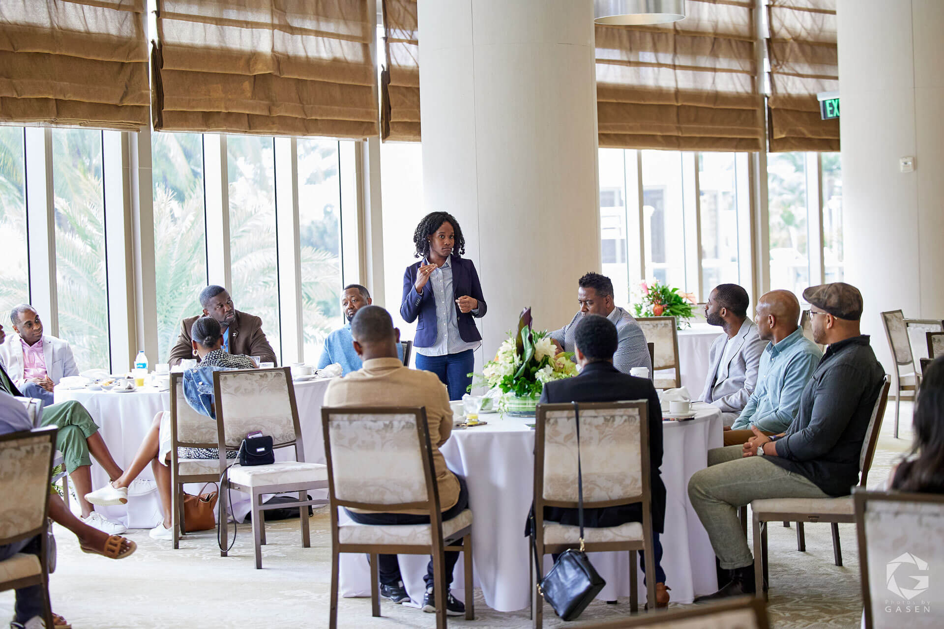 A woman in a navy blazer presenting to an attentive audience in a well-lit conference setting.