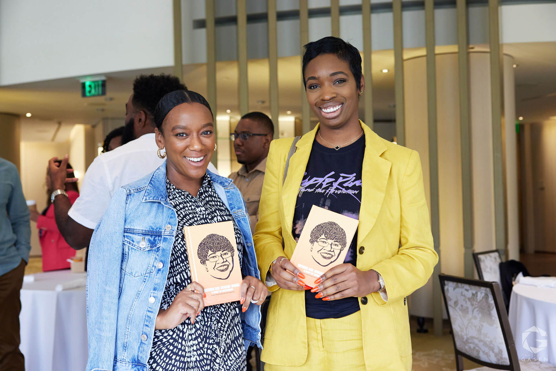 Two smiling women, one in a denim jacket and the other in a yellow blazer, holding books in a casual networking setting.