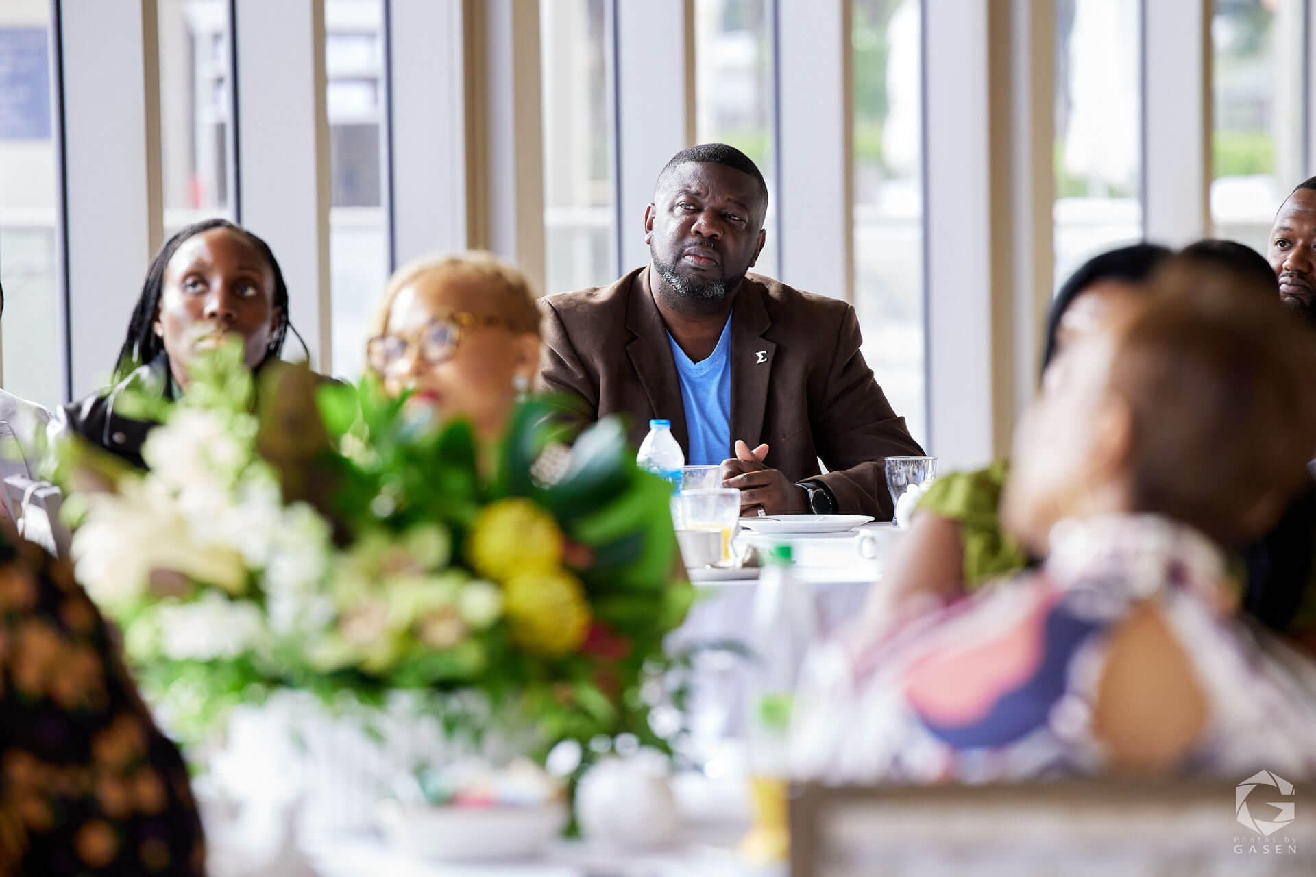A man in a brown blazer with a blue shirt actively listening during a formal gathering, with floral arrangements in the foreground.