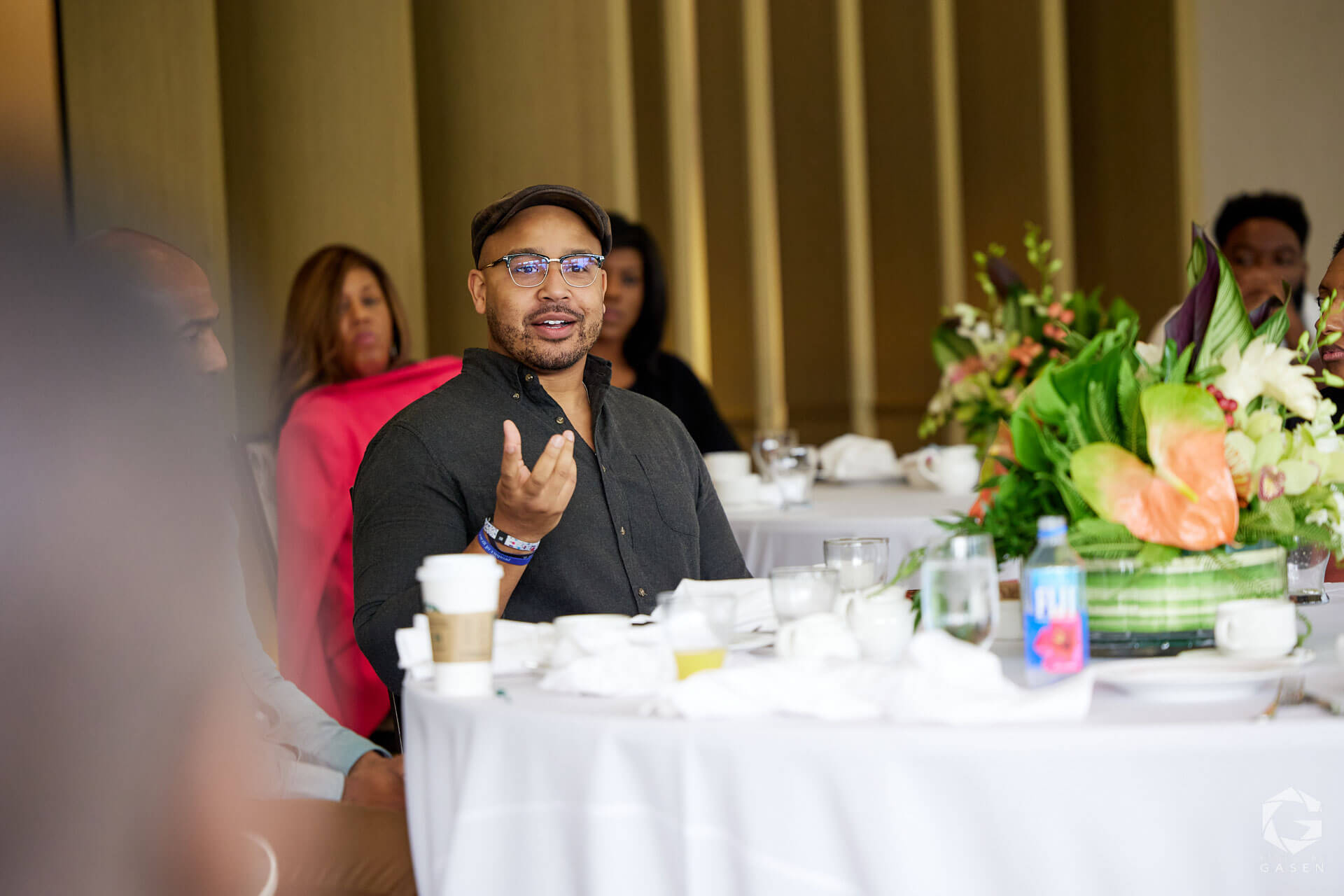 A man wearing glasses and a cap speaking at a table, surrounded by others in a banquet-like setting.