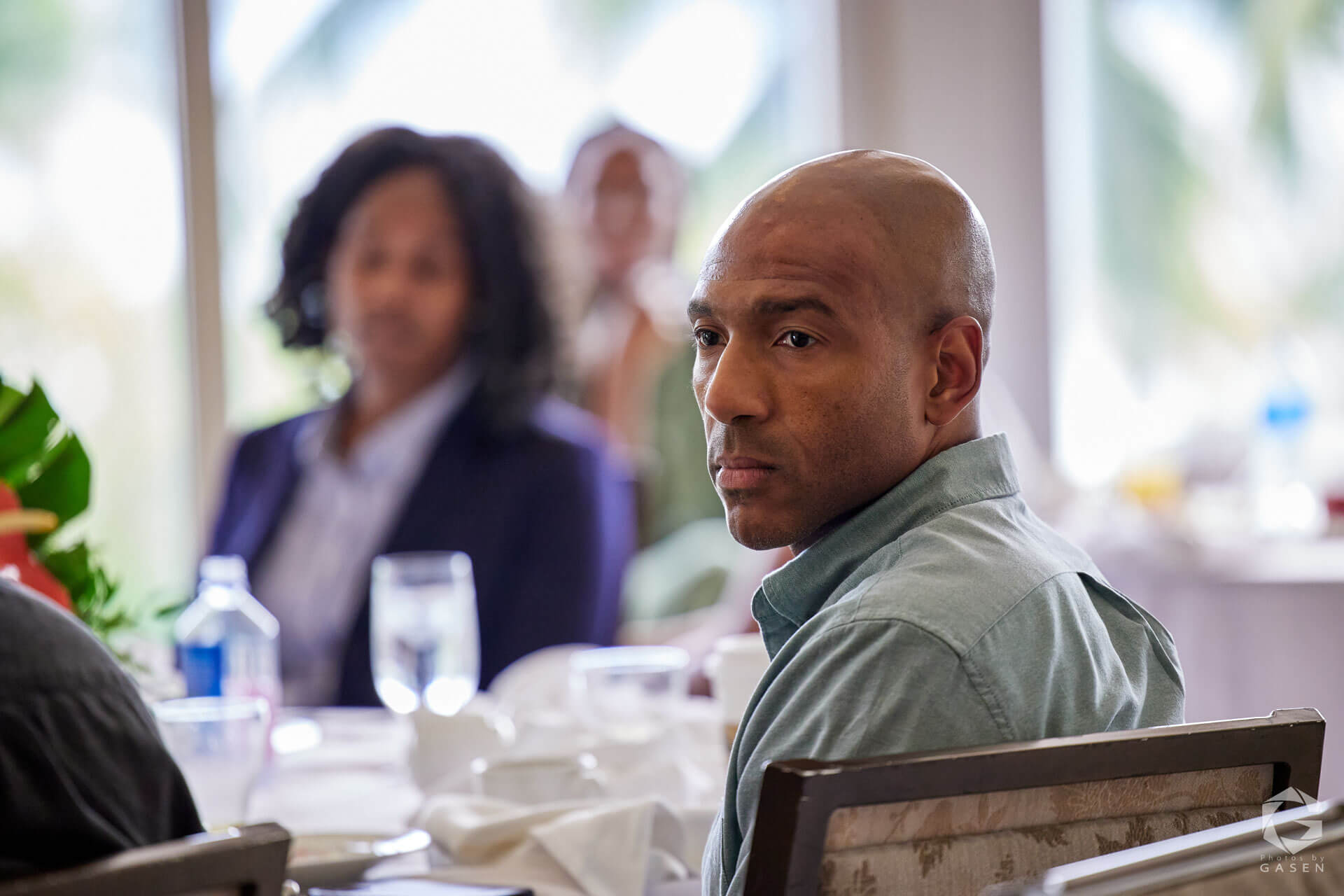 A man seated at a table, looking contemplative with blurred attendees in the background.