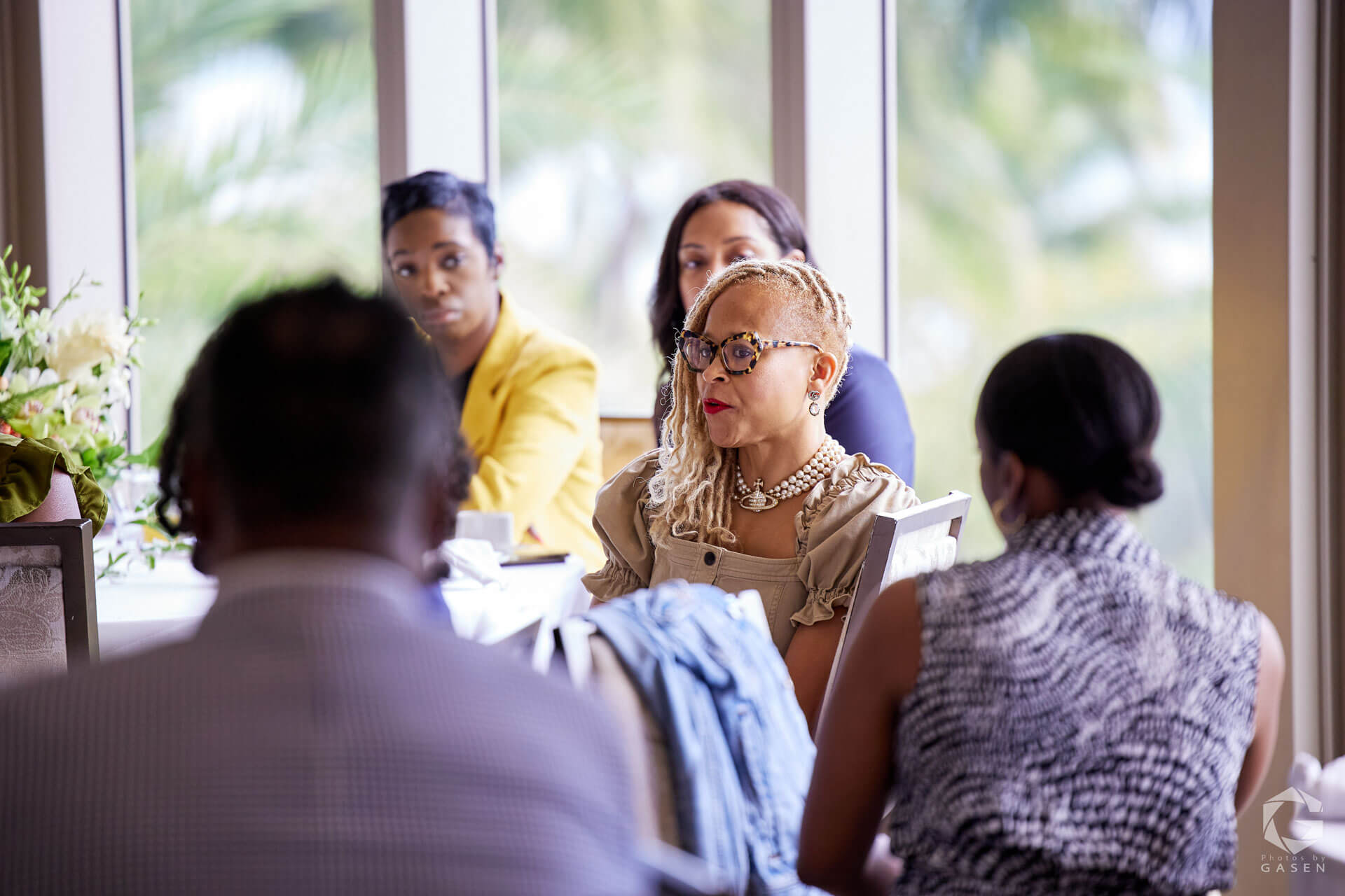 A group of women seated around a table, with a woman in focus wearing a beige outfit and statement glasses.
