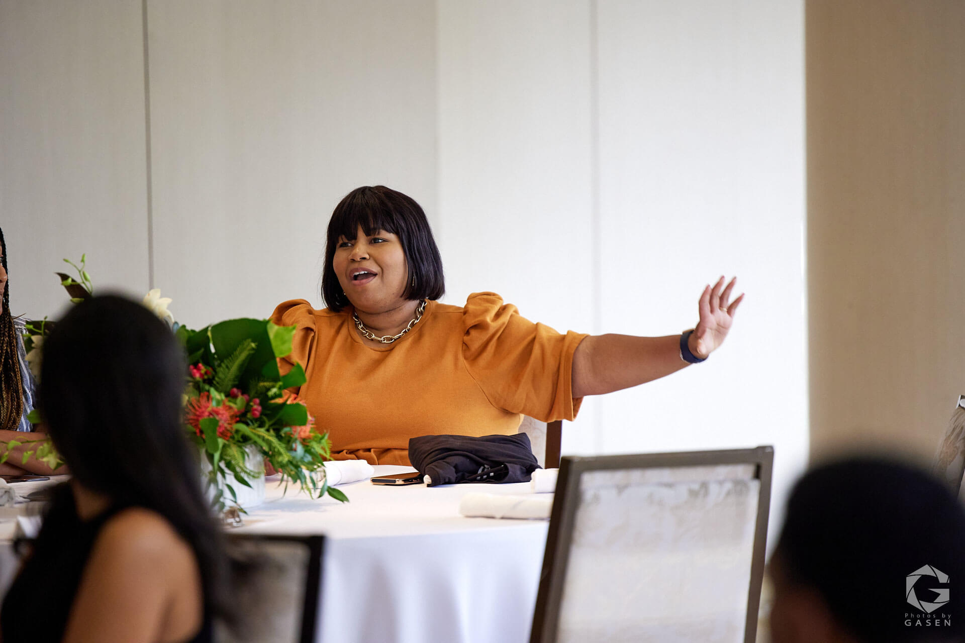 A woman in an orange top animatedly speaking at a table, gesturing with her arms open wide.
