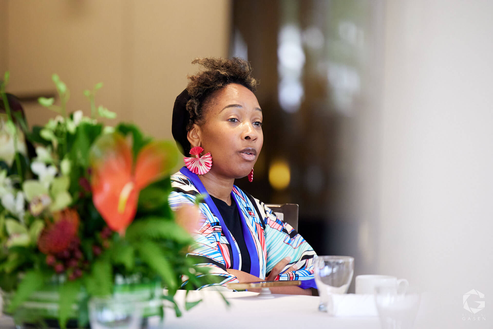 A woman seated at a table with vibrant floral arrangements in the foreground, speaking confidently during a formal event.