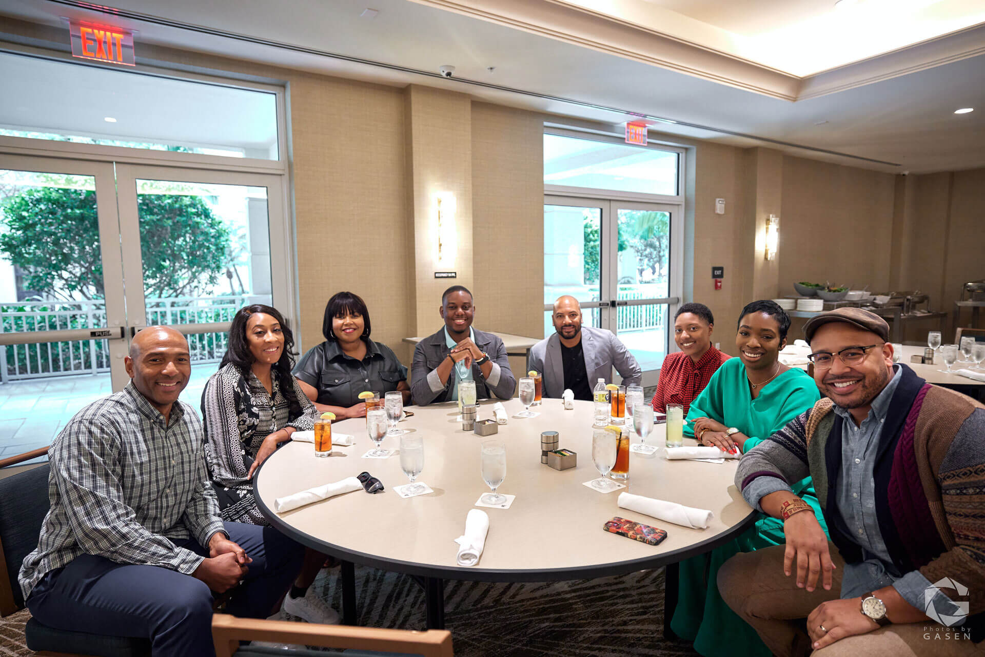 A group of smiling people seated around a round table in a bright conference room, engaged in conversation during a meeting or event.