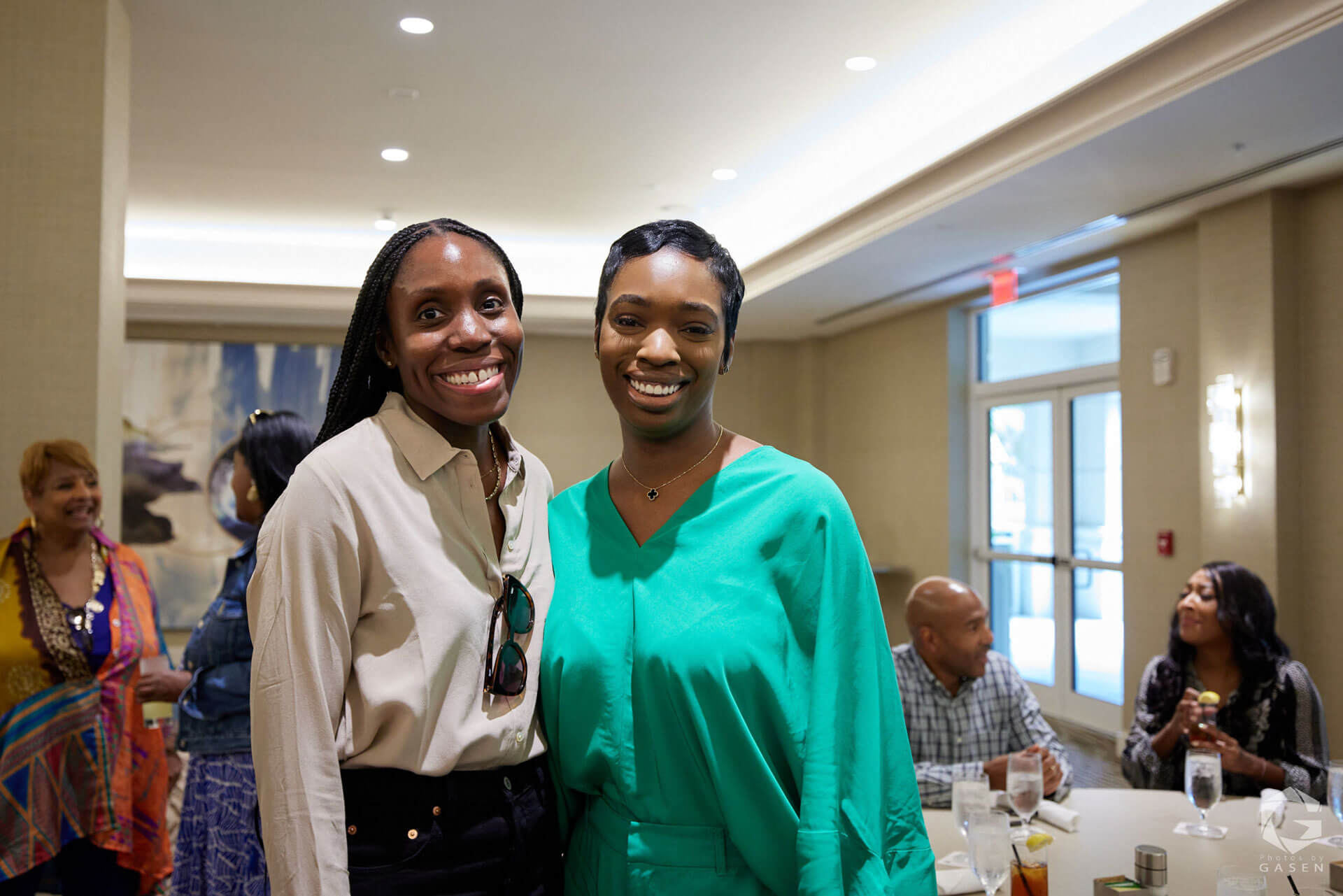 Two women standing together, smiling warmly, with a group of people in the background at an indoor gathering.